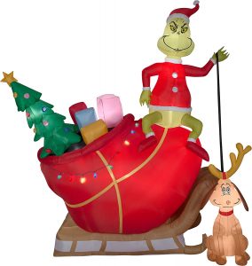An inflatable version of the grinch sitting on top of a sleigh with a red bag full of decorations and presents he stole from the Whos,he is holding a leash attached to the dog with reindeer antlers on